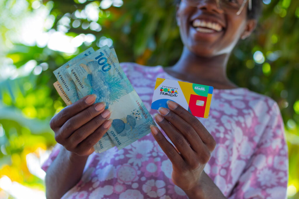 Imagem de mulher sorrindo com cartão do programa Bolsa Família em uma mão e o dinheiro do benefício em outra